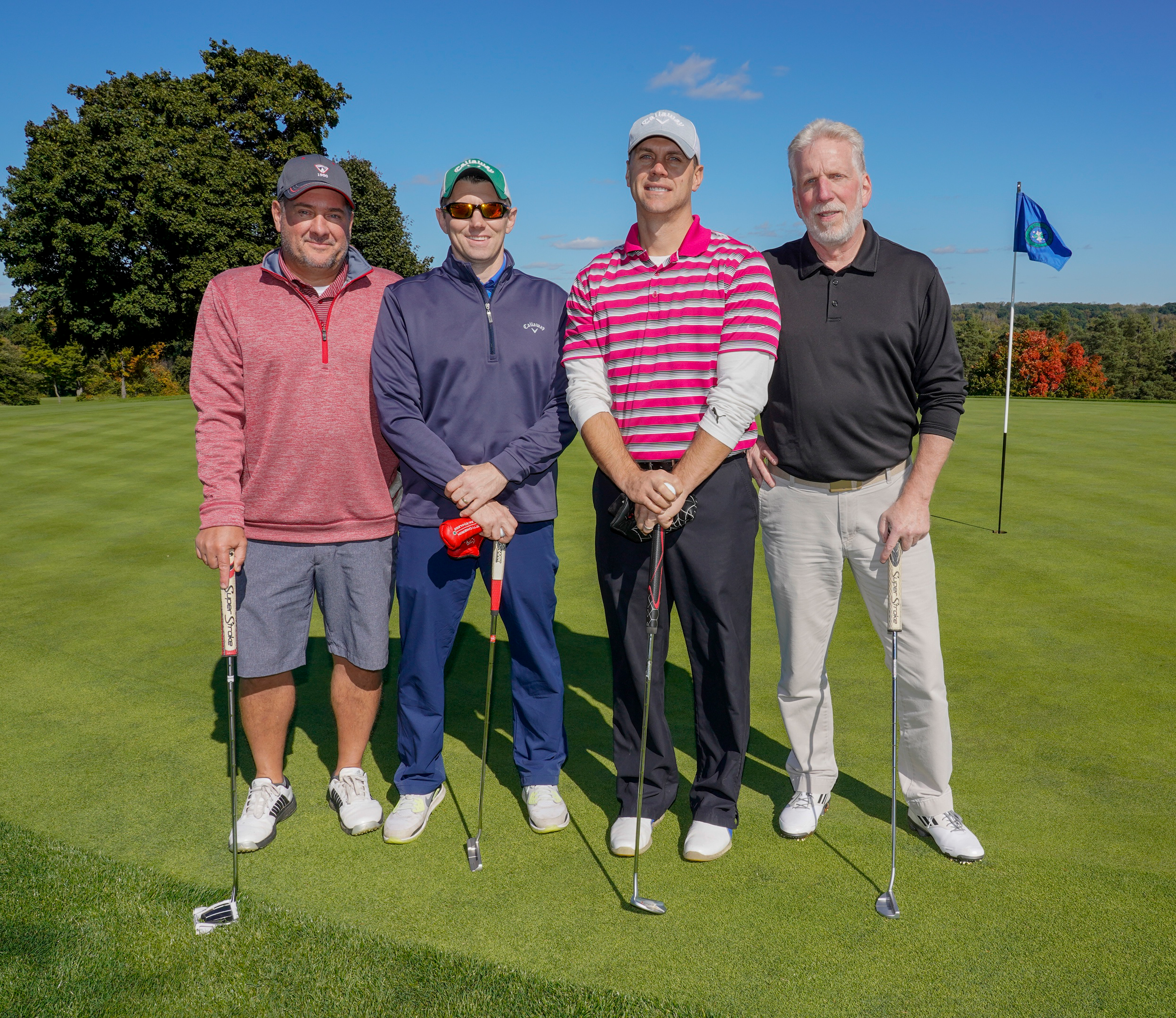 Four men wearing hats stand on a golf course. One holds a golf club.